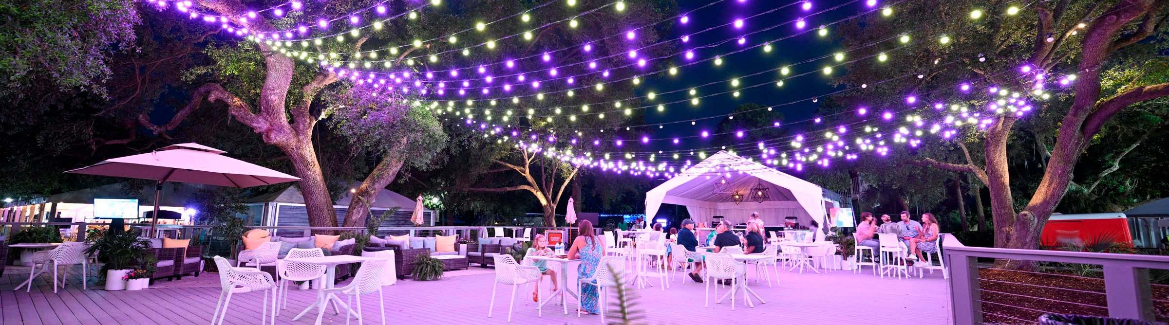 People mingle at tables and chairs beneath a colorful array of string lights during an evening outdoor event. 
