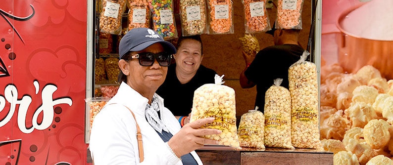 A woman smiling as she holds a bag of popcorn in front of a popcorn vendor.
