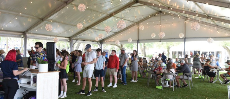 People stand in line on a lush green lawn beneath a large event tent. String lights and decor decorate the roof of the tent. 